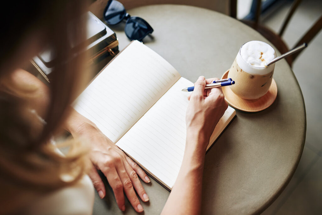 Woman journaling near iced coffee. Recovering from complex PTSD and trauma takes time. Why not speak with a trauma therapist who understands the effects of trauma. Find a therapist near me in our counseling directory for more support. Miami, FL 33131 |Newark, NJ 07102 | Jersey City, NJ 07307