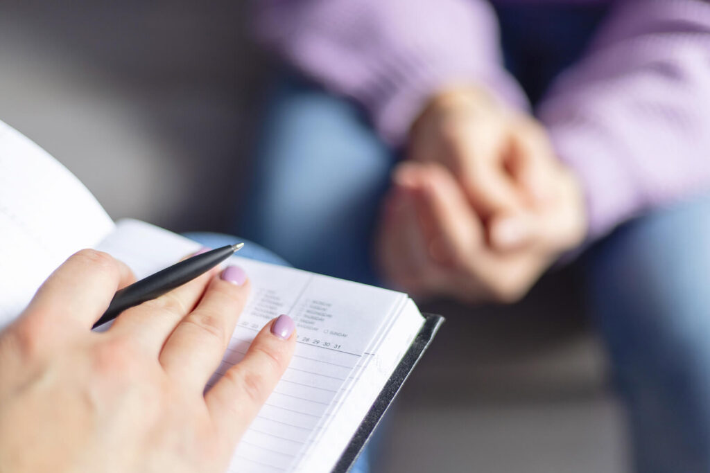 Photo of a person with a notebook and pen sitting across from a woman. Learn how a trauma therapist can offer support in understanding childhood trauma by searching “find a trauma therapist near me” today. They can offer support in healing from childhood trauma.